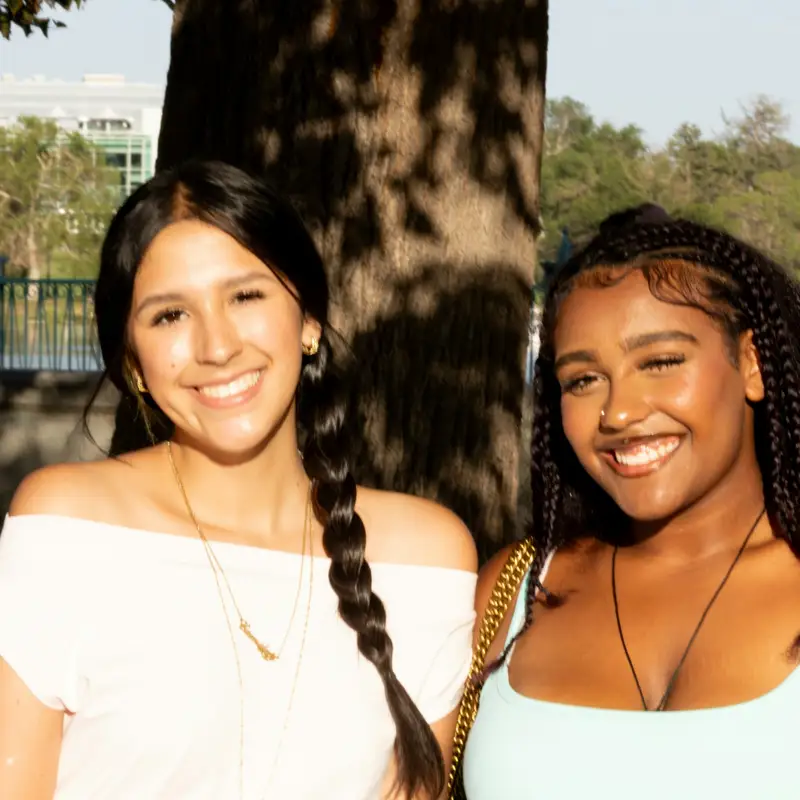 Email header photo: two smiling young women standing together in front of a tree in a sunlit park