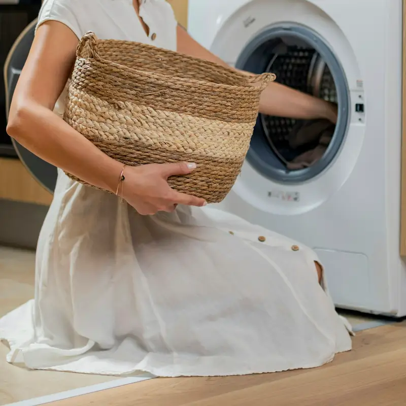 Email header photo: a woman in a white dress kneeling with a woven basket while loading laundry into a front-load washer