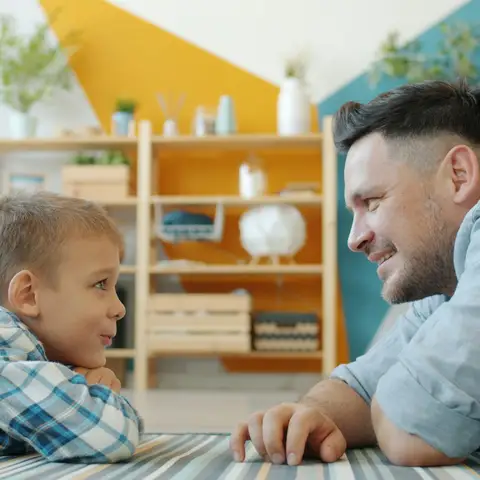 A father and his young son lying on a striped rug in a bright living room, smiling at each other