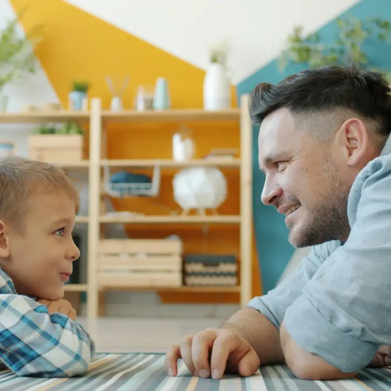 Email header photo: a father and his young son lying on a striped rug in a bright living room, smiling at each other