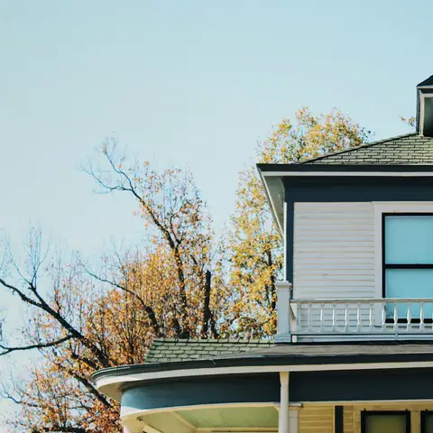 A two-story white house with dark trim, a wraparound porch and autumn trees against a clear sky