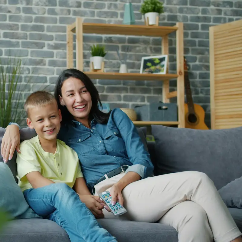 Email header photo: a mother and her young son smiling together on a gray sofa in a bright living room with a brick wall and shelves behind them