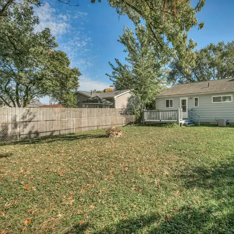 A wide, green backyard lawn behind a single-story gray house with a small deck, a wooden fence and tall shade trees