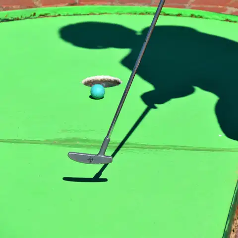 Putter and a blue ball beside the hole on a bright green mini golf course, with the player’s shadow on the turf