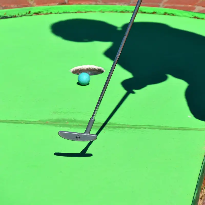 Email header photo: putter and a blue ball beside the hole on a bright green mini golf course, with the player’s shadow on the turf
