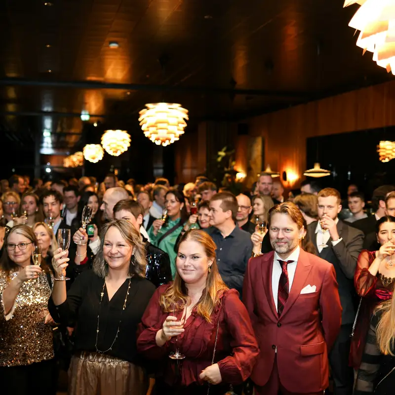 Email header photo: a crowd of guests in evening wear raising their glasses under warm pendant lights at a company celebration