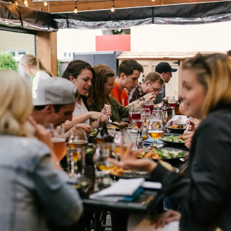 Email header photo: friends talking and sharing plates of food at a long table under string lights during a private party on a covered patio