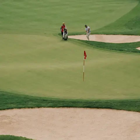 Golfer chipping from beside a sand bunker toward a red flag on a wide green, with a partner and golf bag nearby