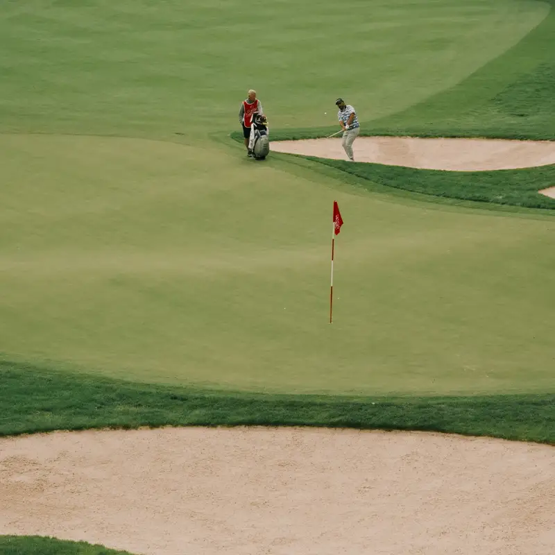 Email header photo: golfer chipping from beside a sand bunker toward a red flag on a wide green, with a partner and golf bag nearby