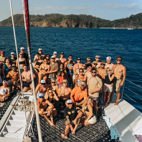Large group of travelers posing together on the deck of a catamaran in a sunny bay