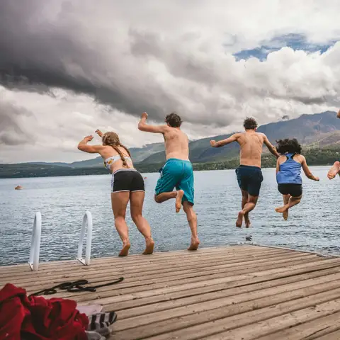Group of friends in swimwear jumping together off a wooden dock into a mountain lake under a cloudy sky