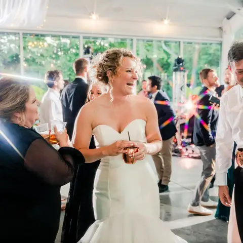 A bride laughing with her guests on a crowded dance floor under string lights at her wedding reception