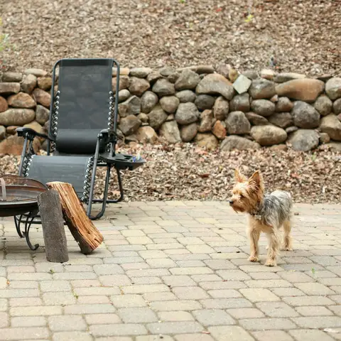 Two reclining chairs around a metal fire pit with stacked firewood on a tan brick paver patio, below a dry-stacked river stone retaining wall, with a small terrier standing nearby