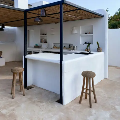 A white plastered outdoor kitchen with a bar counter, sink and open shelves under a dark steel pergola with a reed roof, with two wooden bar stools in front