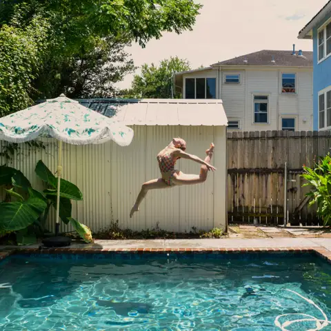 A person leaping through the air above a small backyard pool with a stone edge, a garden shed and an umbrella