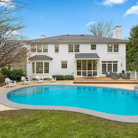 A clear blue backyard pool and spa behind a white two-story house with a deck and lounge chairs