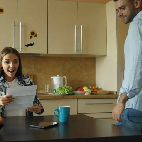 A woman at a kitchen table reading a letter with a delighted look while her partner walks over to see it