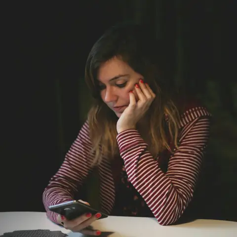 A worried young woman resting her head on her hand while holding a phone at a table in a dark room