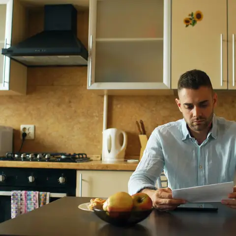 A man in a light blue shirt reading a letter at his kitchen table, with a mug and a bowl of fruit beside him