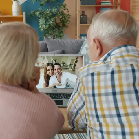 An older couple seen from behind on a video call with a smiling younger couple on the laptop screen
