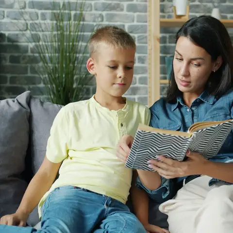 A mother and her young son reading a book together on a gray couch at home