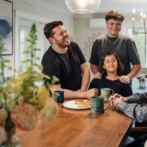 A father, a teenager and a young girl smiling around a wooden dining table with mugs and pastries, an older relative seated in the foreground