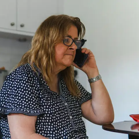 A woman in glasses making a phone call at her kitchen table