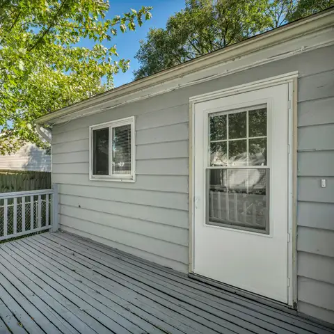 A grey painted wood deck with railings beside a house with grey lap siding, under shade trees