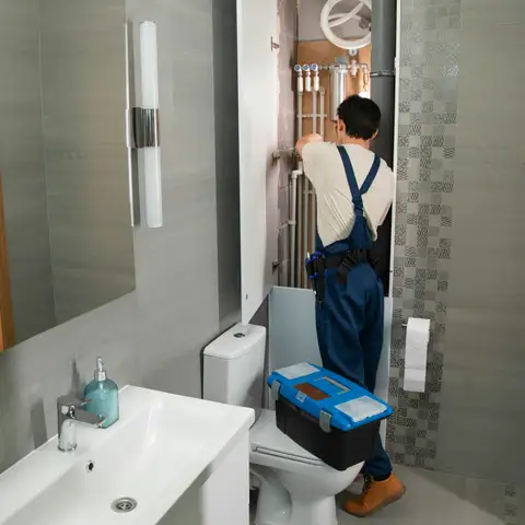 A plumbing contractor in overalls fitting supply pipes inside an open bathroom wall, with a toolbox set on the toilet lid