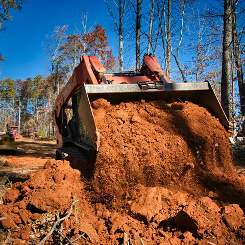 A tracked skid steer loader pushes a full bucket of red clay across a cleared lot in an autumn forest, with a crew member and a second machine behind it