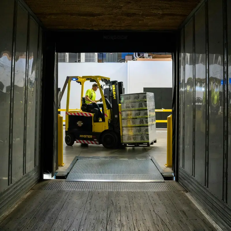 Email header photo: seen from inside an empty trailer, a forklift driver lifts a wrapped pallet of cases toward the open trailer door at a warehouse dock