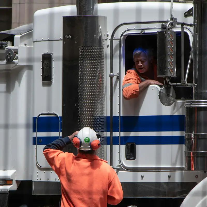 Email header photo: a truck driver leaning out of the cab window of a white tractor to talk with a yard worker in a hard hat and orange high-visibility shirt