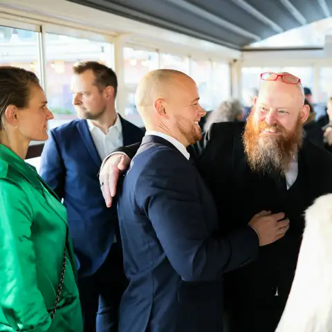 Two smiling men greeting each other with a hug at a crowded evening networking event in a bright glass room
