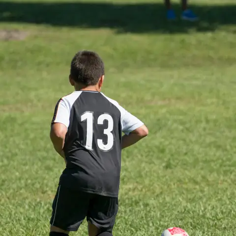 A young soccer player in a black and white number 13 jersey seen from behind, hands on hips, next to a ball on the grass