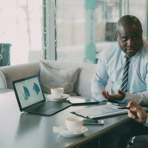 A man in a shirt and tie talking with a colleague over coffee, with pie charts on an open laptop and a printed report on the table