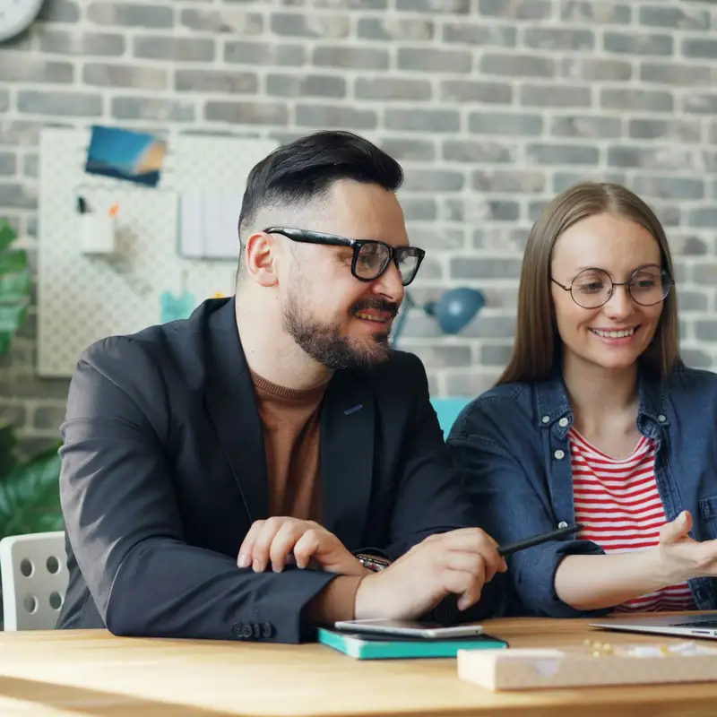 Email header photo: a man in glasses and a woman in a denim jacket smiling at a laptop together at a wooden desk in front of a brick wall