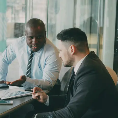 Two men in business clothes sit at an office table reviewing charts on a laptop and printed papers