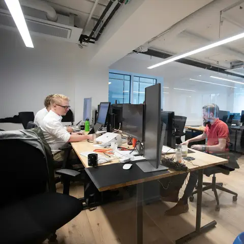 An office team working at rows of desks with large monitors in a bright open-plan office with tall windows