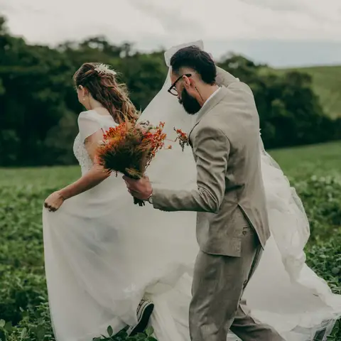 A bride holding a bouquet and a groom in a tan suit running hand in hand through a green field