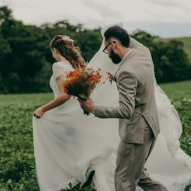 Email header photo: a bride holding a bouquet and a groom in a tan suit running hand in hand through a green field