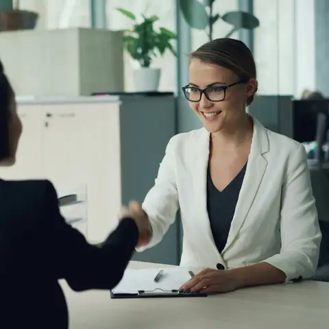 A smiling woman in glasses shaking hands across an office desk with a new candidate