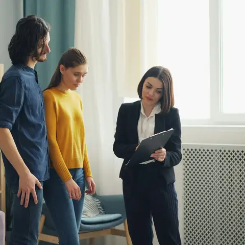A young couple in a bright living room looking over a clipboard with a woman in a dark suit