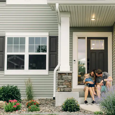 A couple holding their baby on the front steps of a gray-sided home with a white garage door and fresh landscaping