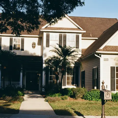 A two-story cream house with a covered front porch, palm trees, a mailbox and a walkway through the front garden