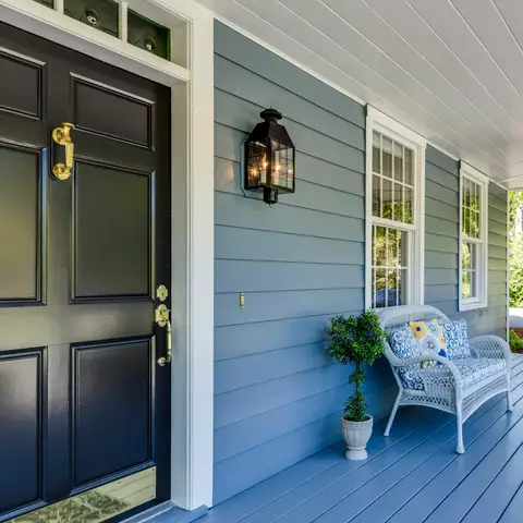A painted blue front porch with a black front door, a brass lantern, a white wicker chair and a hanging porch swing