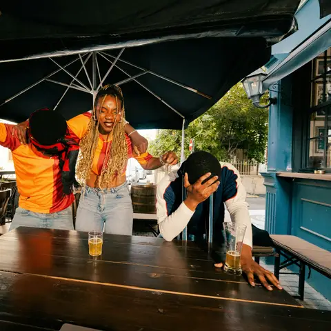 Three friends at a wooden table on a bar patio reacting to a game, one cheering with an arm raised and one holding his head
