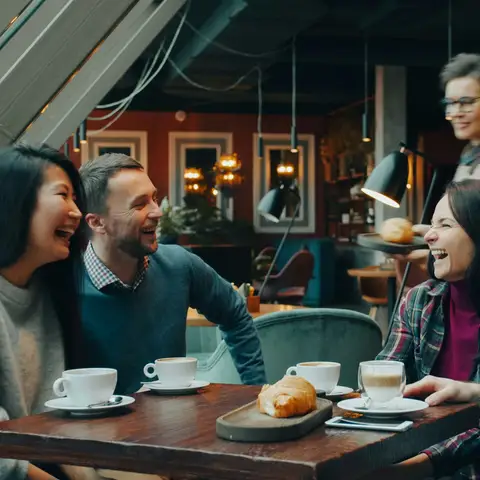 Four friends laughing at a wooden cafe table with cappuccinos and a croissant