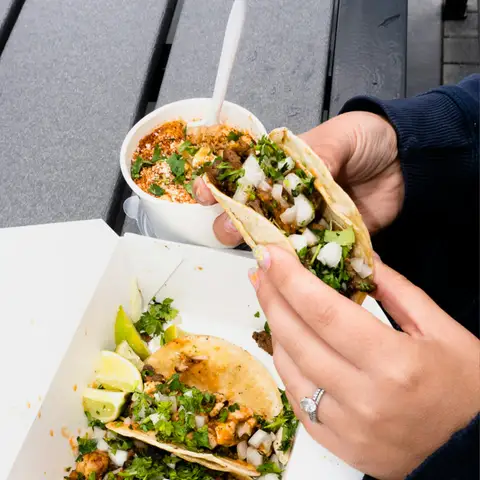 Hands holding a street taco over a box of tacos with lime wedges and a cup of street corn on a picnic table