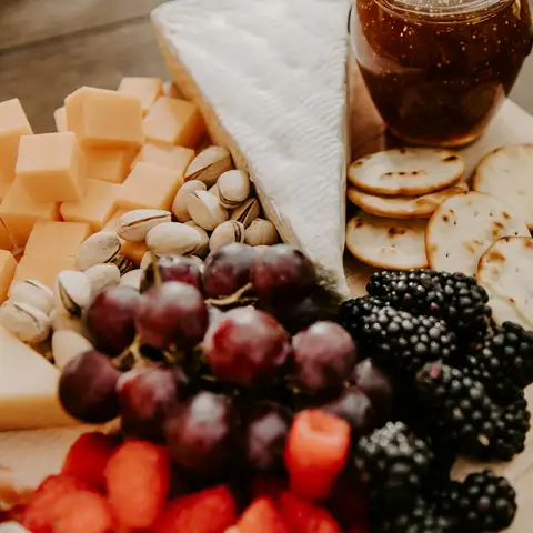 A wooden board with brie, cubed cheddar, pistachios, crackers, grapes, blackberries and a jar of fig jam