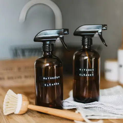 Two amber glass spray bottles labelled bathroom cleaner and kitchen cleaner on a wooden counter beside a dish brush and a cloth